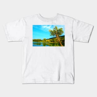 Scene with a green tree hanging above the waters of the lake with vegetation on its shores and Marche hills in the background Kids T-Shirt